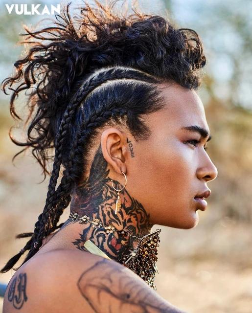 Portrait of an adult male model with braided hair, tiger neck tattoo, and jewelry, gazing right against a soft outdoor background.