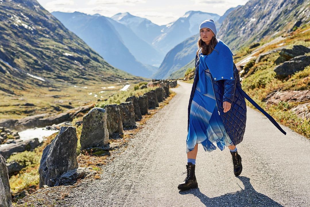 Fashion editorial shot of a stylish woman in a blue dress and puffy coat, walking on a path in a mountain landscape with blue sky.