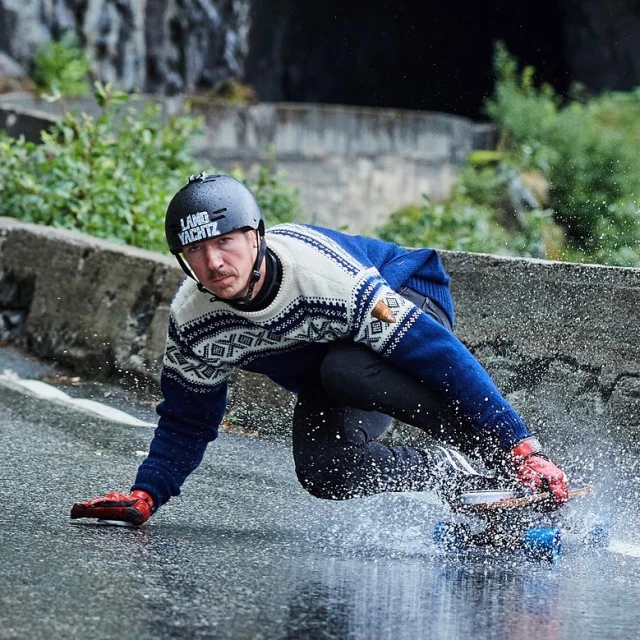 Adult male skateboarder in helmet and sweater speeds down wet road, surrounded by splash of water, concrete barriers, and mountains.