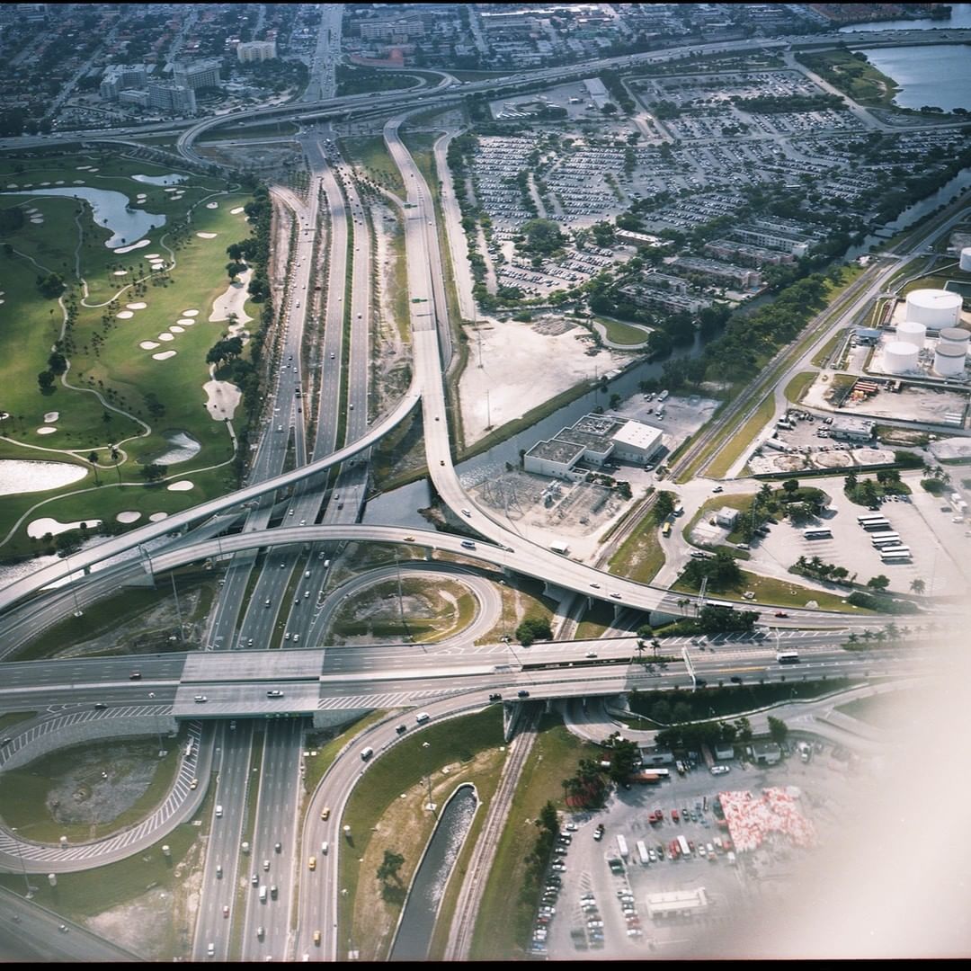 Aerial view of an elaborate highway interchange with multiple overpasses and access roads, surrounded by a golf course, parking lots, and buildings.