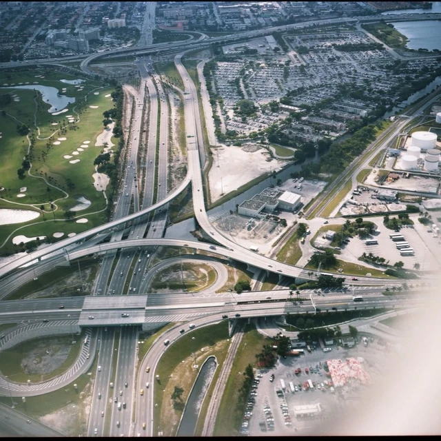 Aerial view of an elaborate highway interchange with multiple overpasses and access roads, surrounded by a golf course, parking lots, and buildings.