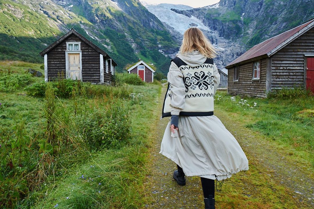 Woman walking on a path near wooden cabins and a glacier-capped mountain range. The scene is set in a lush, green environment.