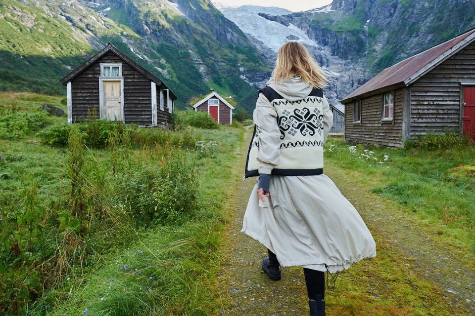 Woman walking on a path near wooden cabins and a glacier-capped mountain range. The scene is set in a lush, green environment.
