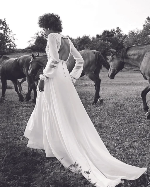 Woman in long white dress in a grassy field with several horses, captured in a monochromatic image with trees in background.