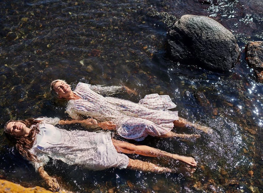Two women in dresses float in shallow water near rocks. Sunlight reflects off the water's surface in an outdoor setting.