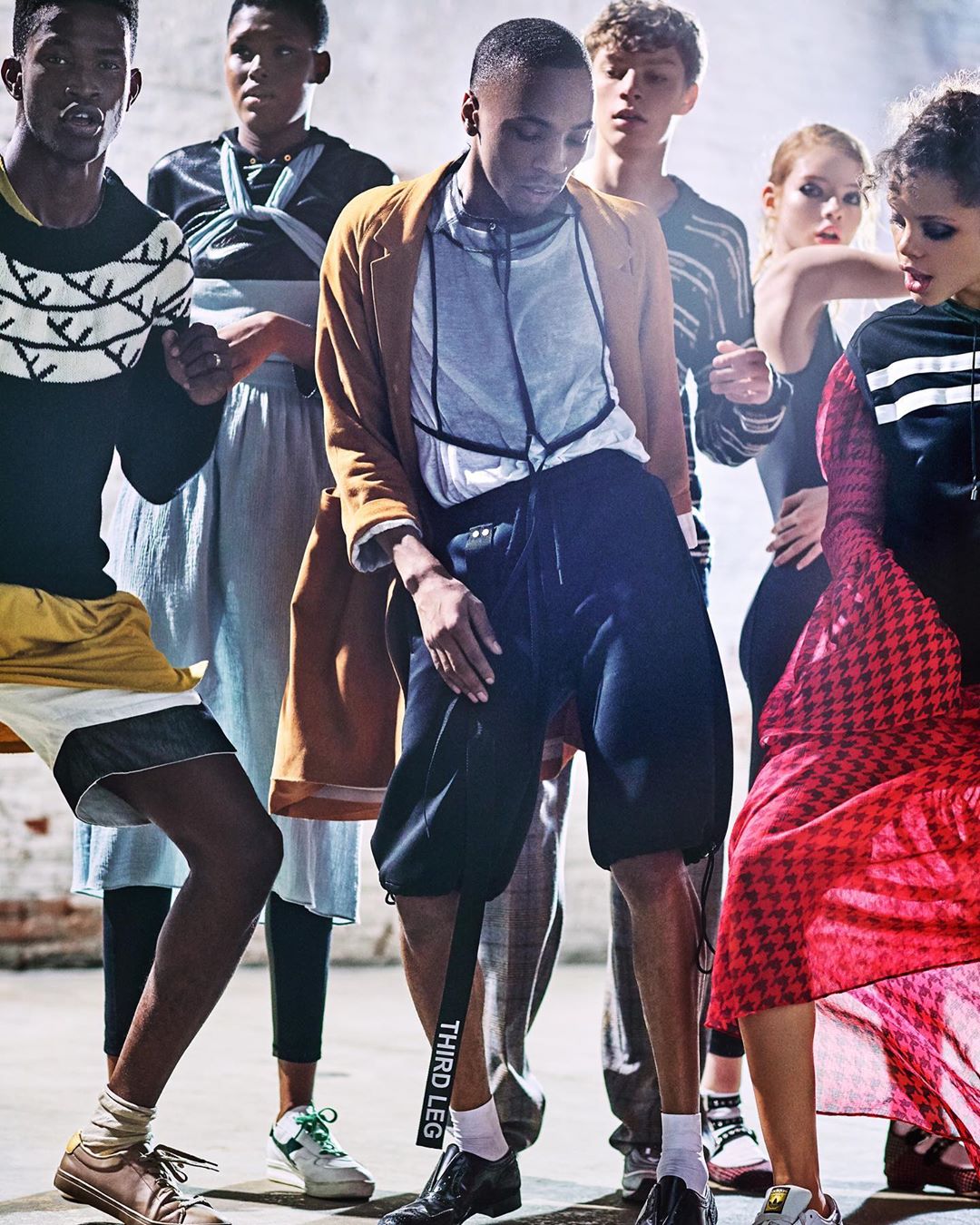 Group of diverse fashion models posing in studio with dramatic lighting, wearing trendy streetwear, blazers, and skirts in a fashion editorial.