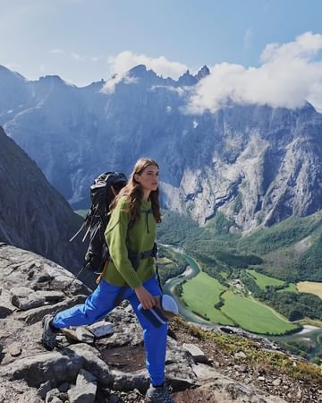 Woman hiking in mountains, wearing a green sweater and blue pants, carrying a large backpack and shoes, with a scenic mountain vista.