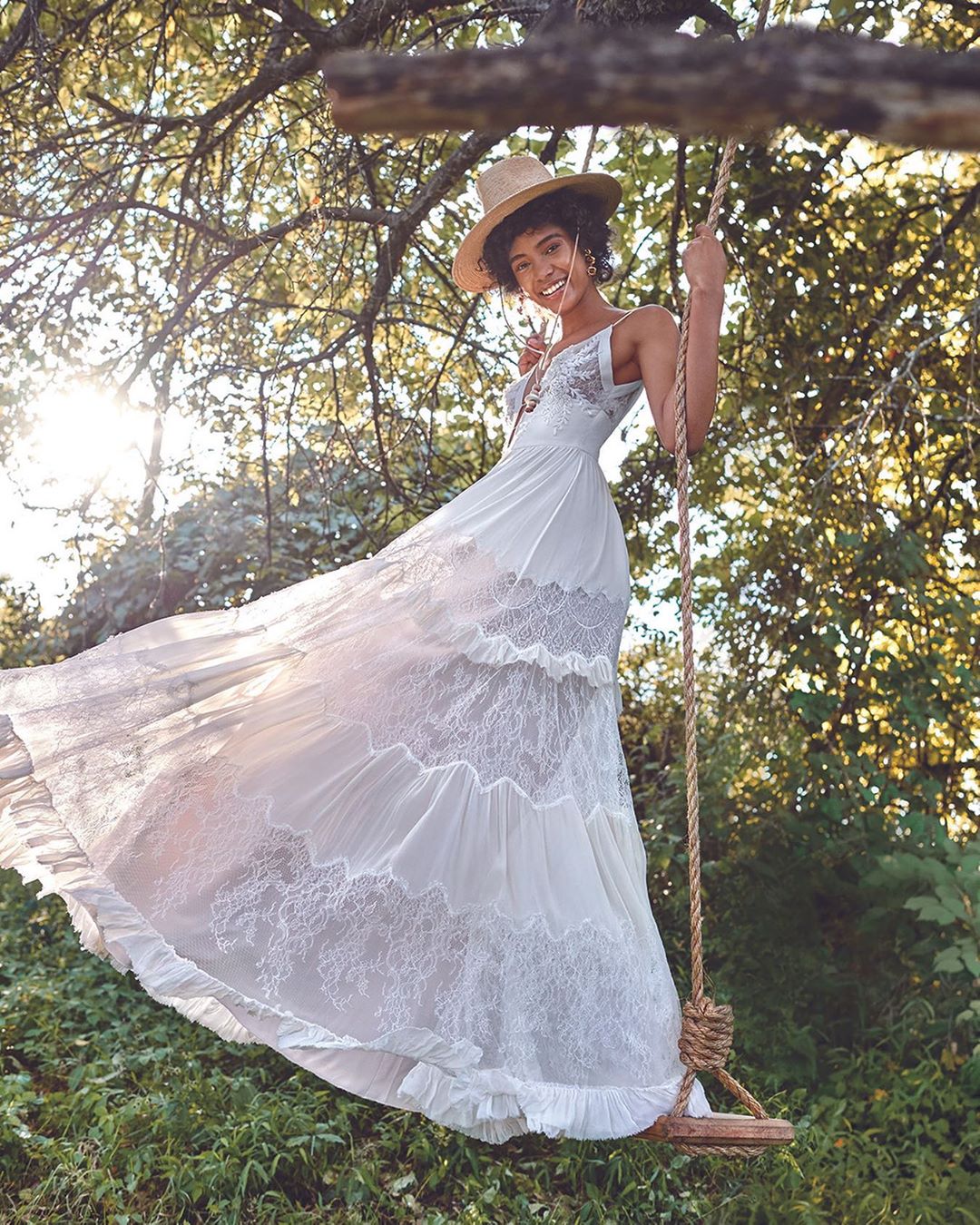 A young woman in a white lace wedding dress and straw hat sits on a swing hanging from a tree in a green, sunny, outdoor setting.