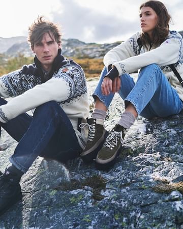 Couple sits on a rock in patterned sweaters and hiking boots against a mountain backdrop, creating an outdoors fashion scene.