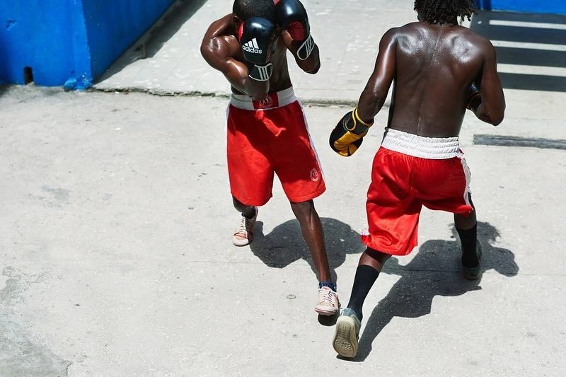 Two muscular men in red boxing shorts and gloves spar outdoors in sunlight with focus on their athletic builds and intense competition.