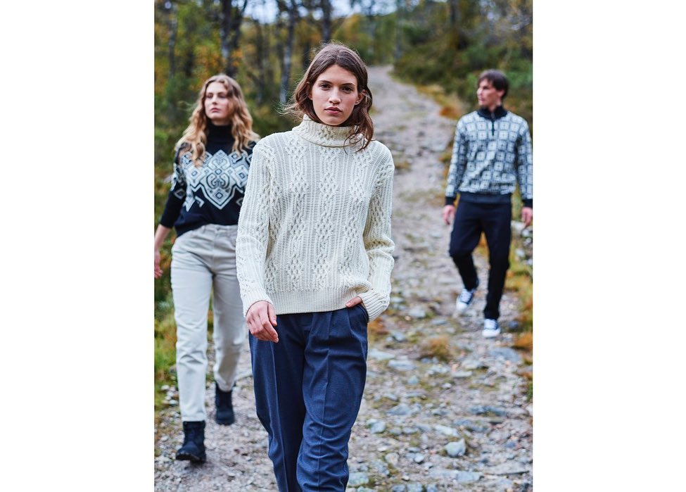 Three adult fashion models pose in sweaters on a rocky path with green trees in the background, wearing pants and walking toward the viewer.