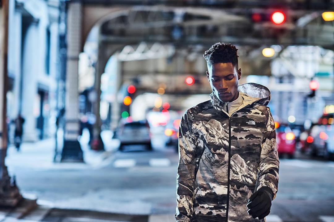 Adult man walking on a city street wearing a grey mountain-patterned jacket and gloves, with street lights and buildings in the background.