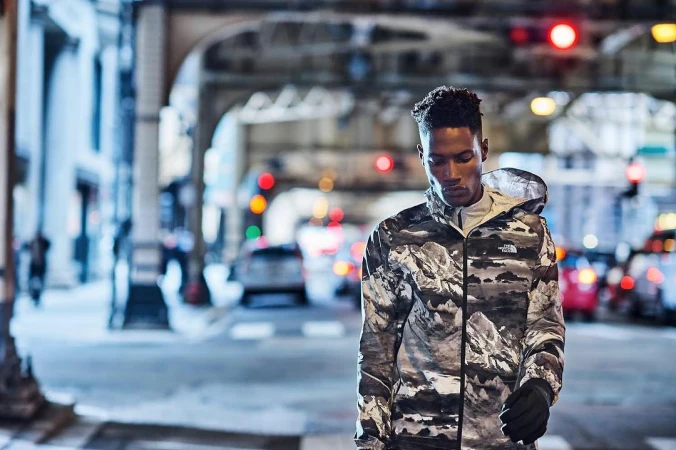 A Black man walking on a New York City street wearing a hooded jacket with a mountain landscape print.