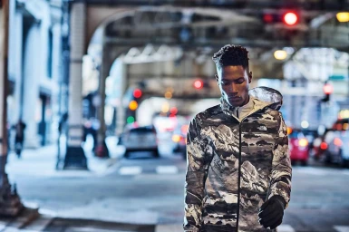 A Black man walking on a New York City street wearing a hooded jacket with a mountain landscape print.