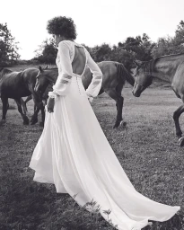 Black and white editorial fashion image of a woman in a long white dress walking outdoors among several horses.