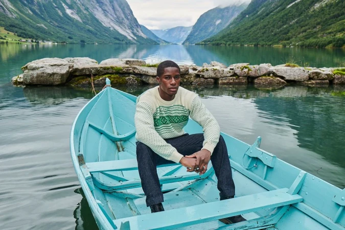 A man seated in a light blue wooden rowing boat on a lake surrounded by steep mountains.