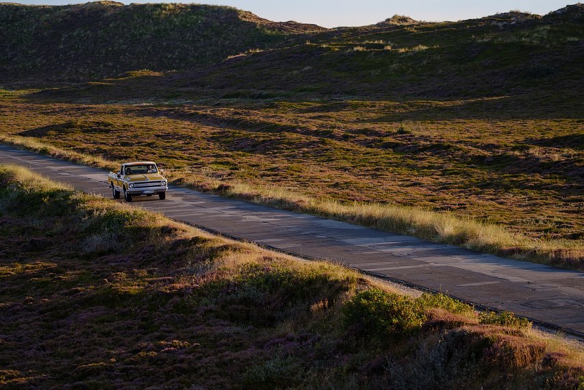 Vintage yellow pickup truck drives down a paved road through a grassy, hilly landscape under a clear blue sky at golden hour.