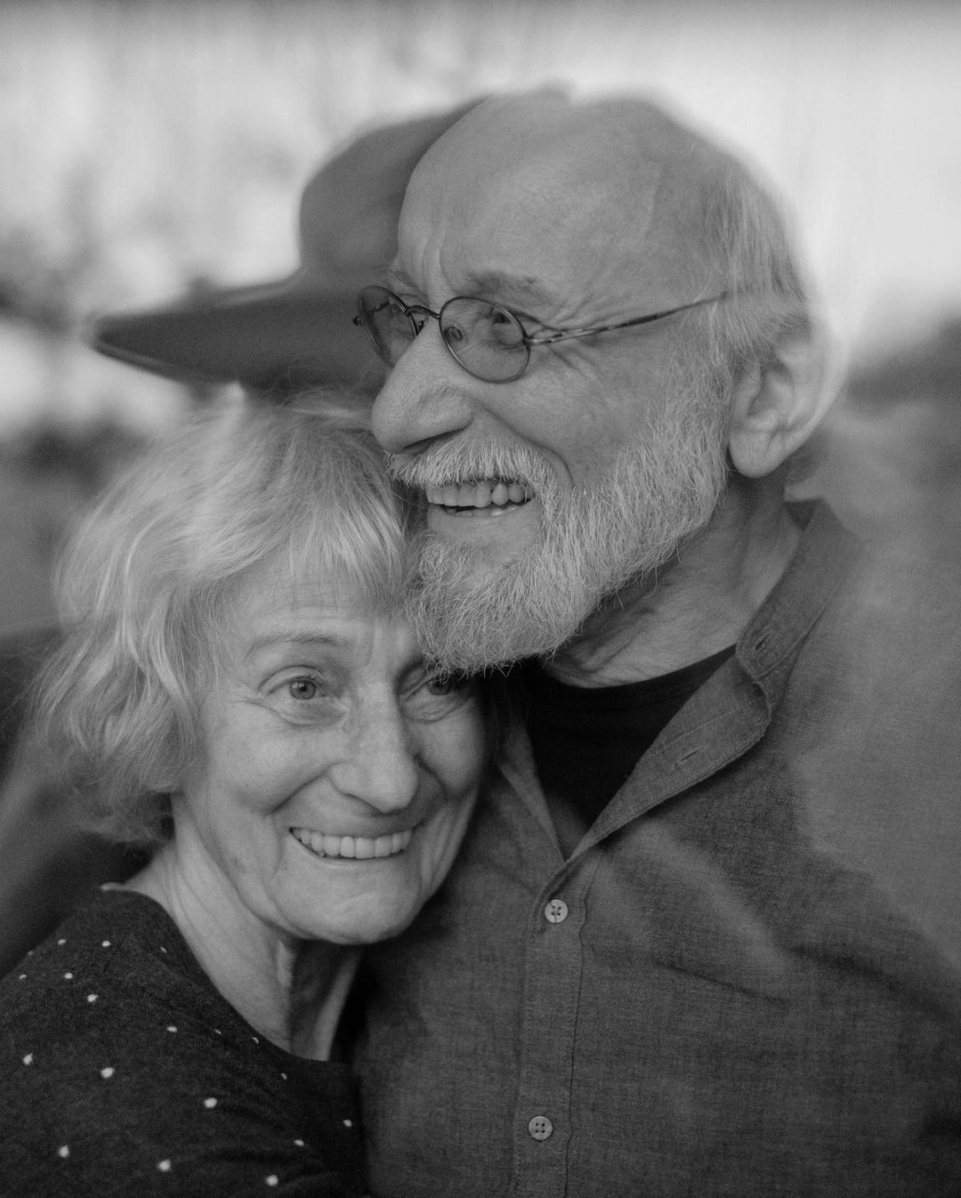Elderly couple embraces outdoors in black and white. The man has glasses and a beard and the woman has a polka dot shirt. Both are smiling.