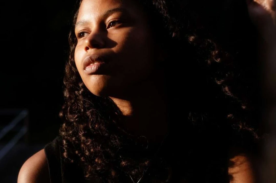 Portrait of a young Black woman with curly hair, bathed in warm light against a dark background, emphasizing her facial features.