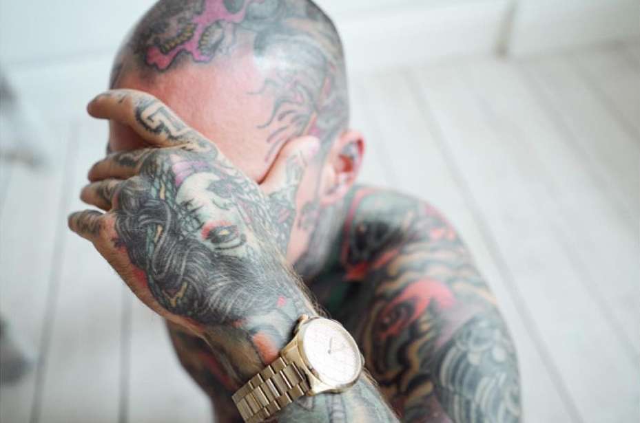 Close-up of a tattooed person covering their face with a hand, wearing a gold watch indoors, with white wood flooring.