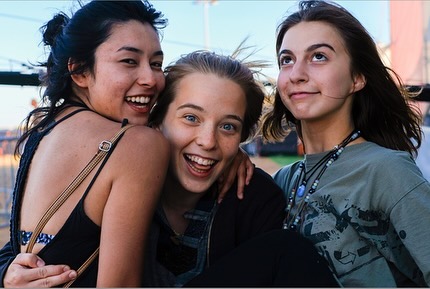 Three happy young women smile and embrace outside, two looking at the camera, one gazing up to the right in an eye-level medium shot.