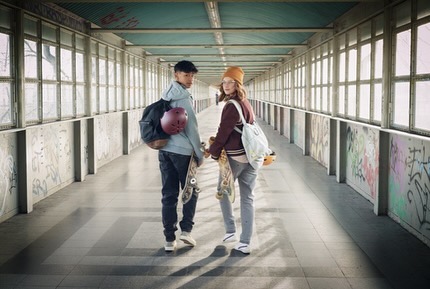 Teenagers with skateboards in an urban tunnel: A boy with a helmet and a girl in a beanie pose with their boards, backlit.