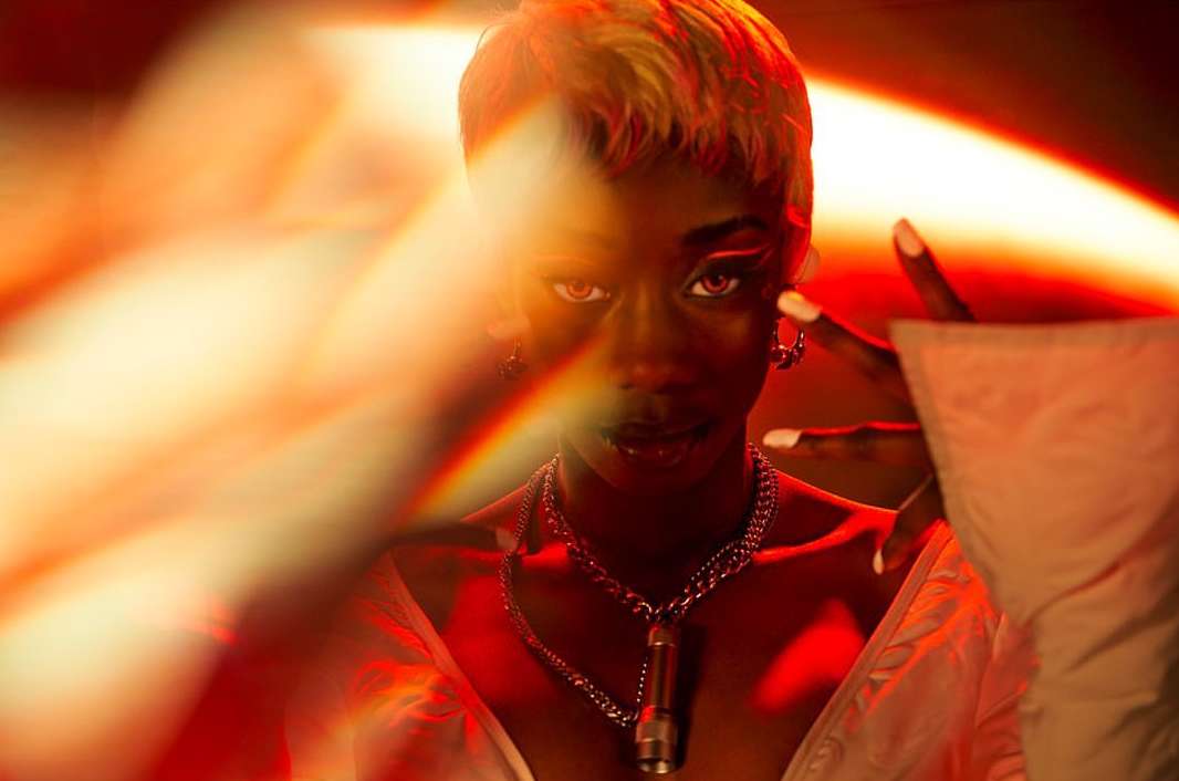 Close-up of a Black woman with short blonde hair and heavy makeup, wearing jewelry, shot with orange lighting and foreground blur.