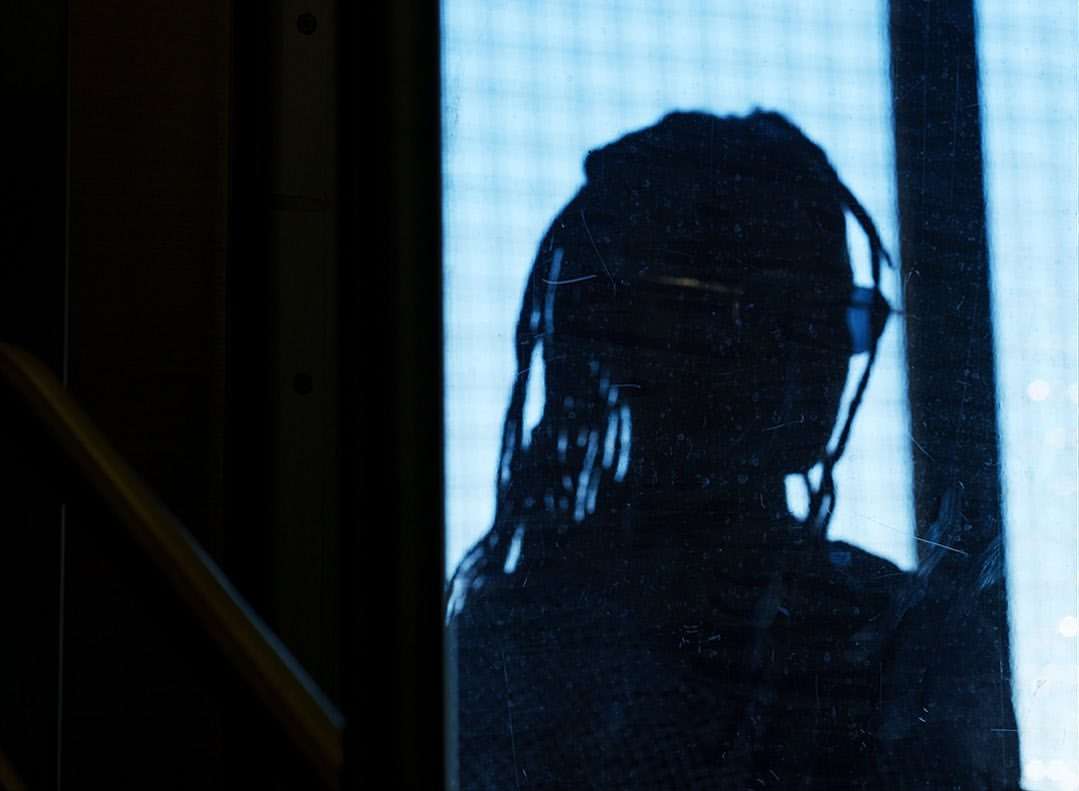 Silhouette of a person with dreadlocks wearing glasses, against a softly lit, gridded background and dark foreground.