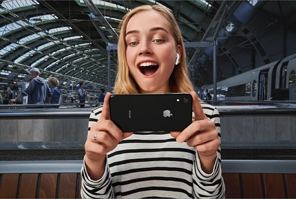 Blonde woman smiles at smartphone in train station wearing striped shirt and white wireless earbuds, passengers in background.