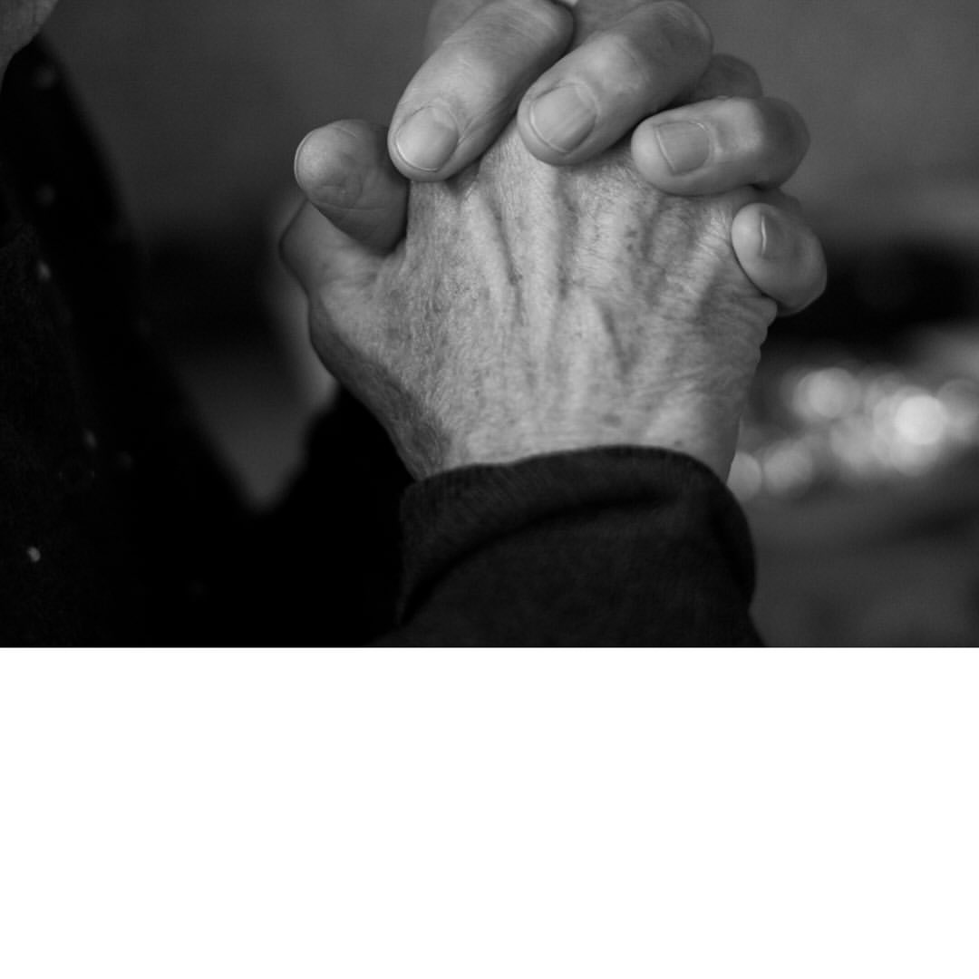 Close-up black and white image of an elderly person's hands clasped tightly, showing wrinkled skin and prominent veins against a blurred background.