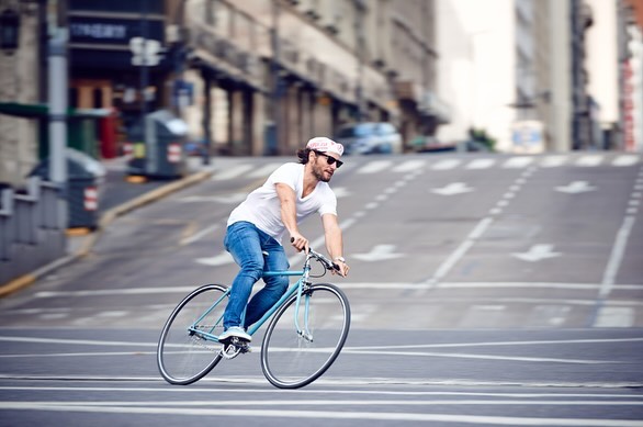 Man riding a blue single-speed bike on city street with buildings, crosswalk, and upward slope, wearing jeans, t-shirt, hat, and sunglasses.