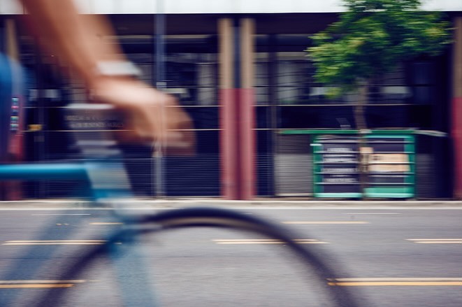 Motion blur photo of a person riding a blue bicycle on a city street with buildings and a tree in the background, capturing urban movement.