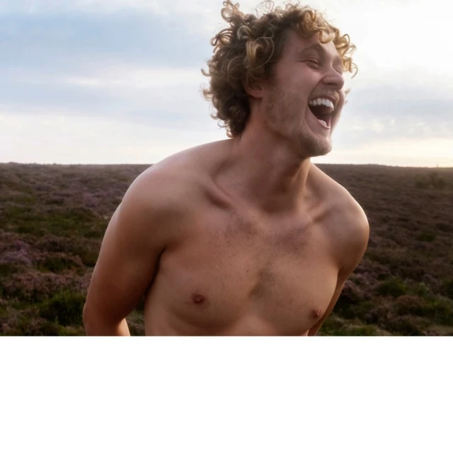 Shirtless adult male with curly blonde hair laughs in an outdoor field setting, showing positive emotion in a natural light photo.