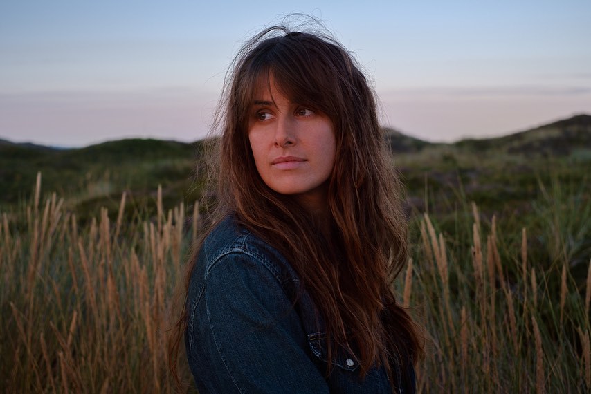 Woman with long brown hair stands in tall grass with a denim jacket, looking over her shoulder at dusk in a natural setting.