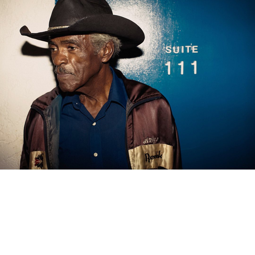 Portrait of an elderly African-American man in a cowboy hat and jacket, standing in front of a blue wall with white lettering.