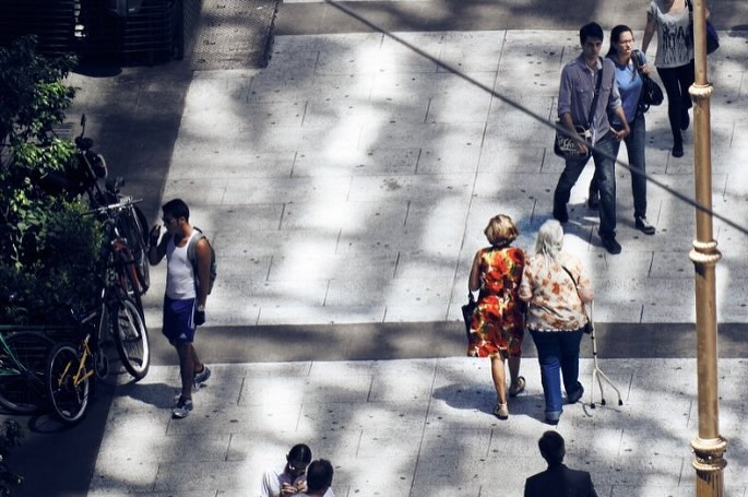 Overhead view of pedestrians walking along a grey-tiled urban walkway with dappled tree shadows on a bright, sunny day.