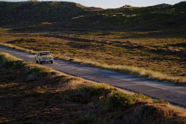Vintage yellow pickup truck driving along a narrow asphalt road through a hilly moorland landscape at sunset