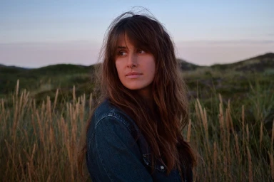 Portrait of a woman with long brown hair wearing a denim jacket outdoors amid tall grass at dusk by photographer Cecil Arp.