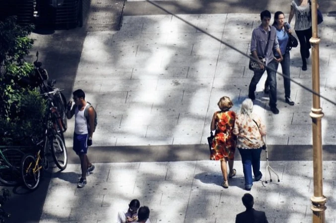 High-angle street photography showing pedestrians walking on a tiled pavement with dappled sunlight and parked bicycles.