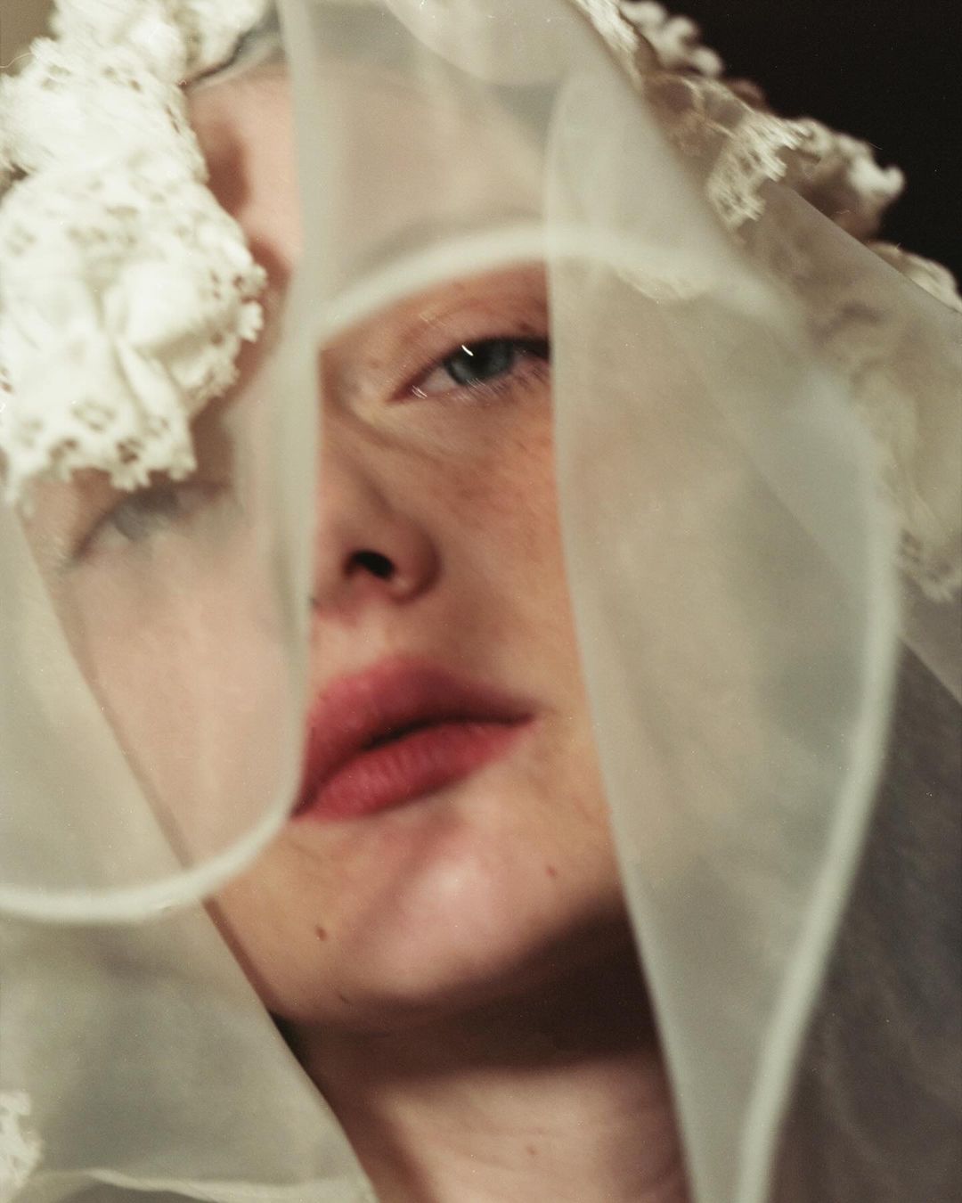 Close-up portrait of a fair-skinned adult female with freckles, wearing a sheer white head covering with lace trim, softly lit.