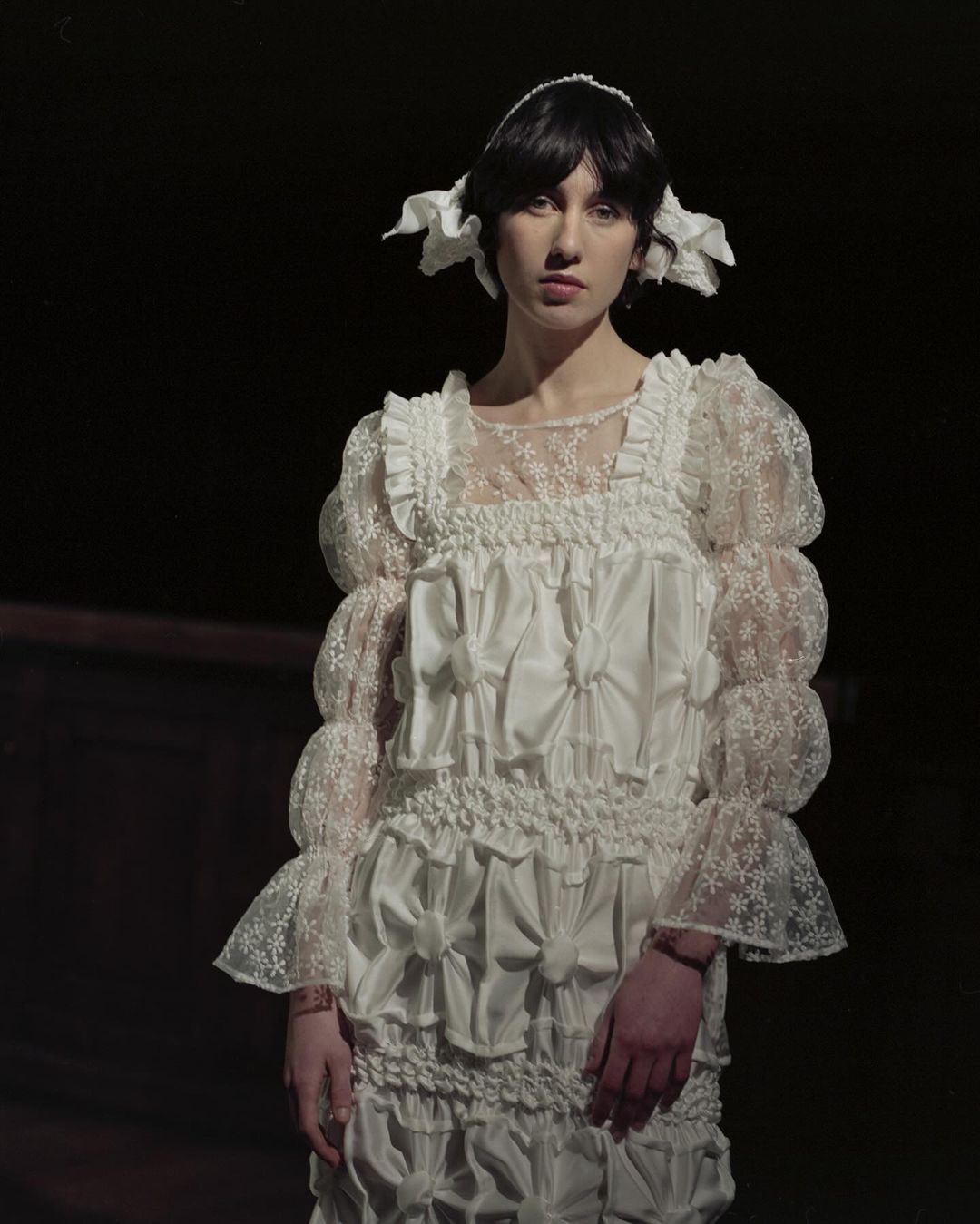 Portrait of a young woman wearing a white lace dress with puffy sleeves and bows against a dark background in a studio setting.
