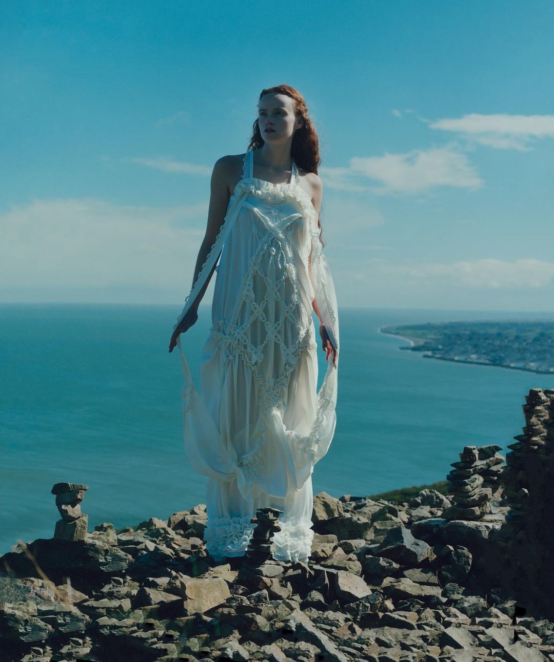 A woman in a white gown stands on a rocky clifftop, ocean and skyline in the background, creating a fashion editorial photograph.