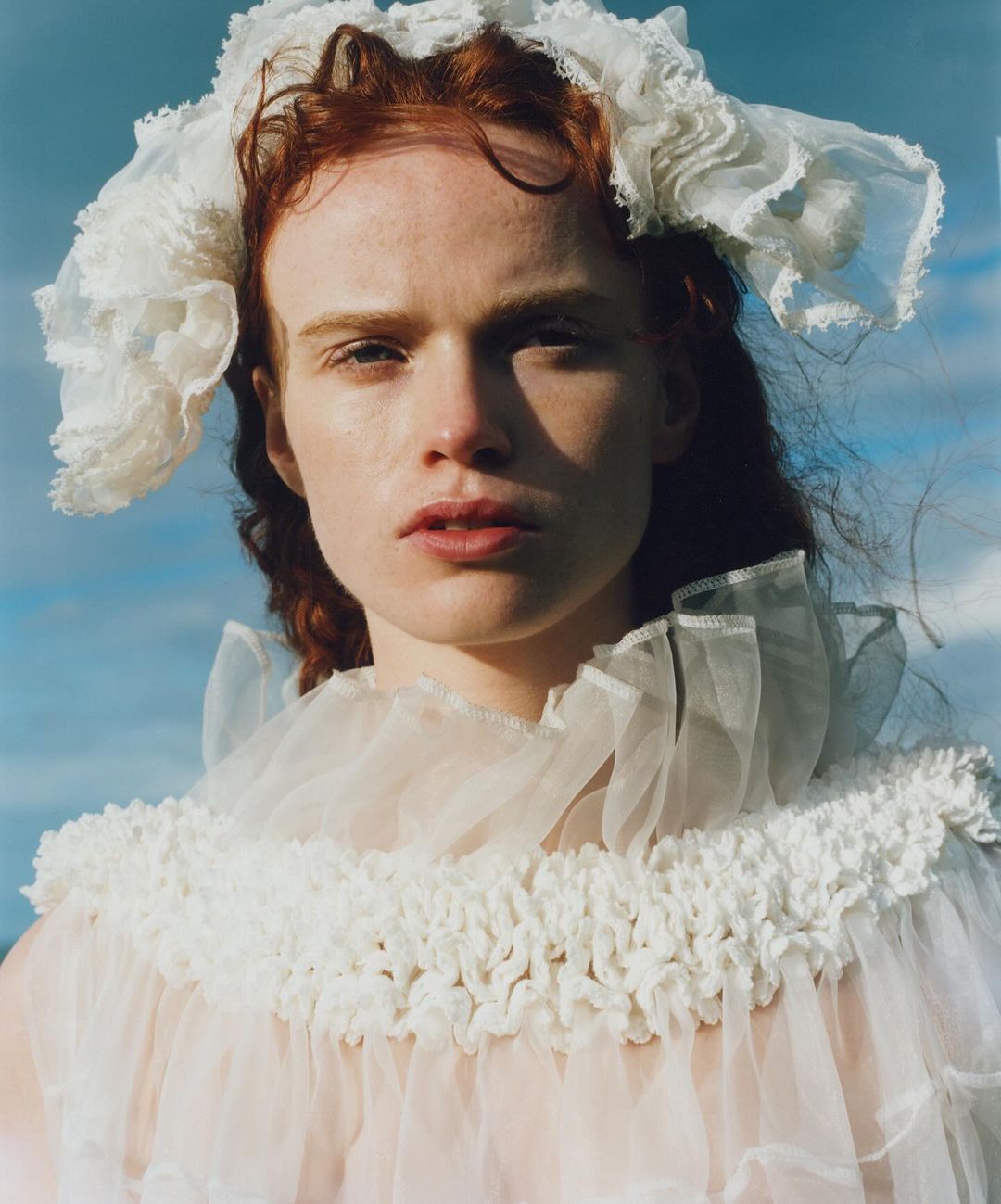 Portrait of an adult female with red hair wearing a sheer white frilled dress and bonnet against a light blue sky with clouds.