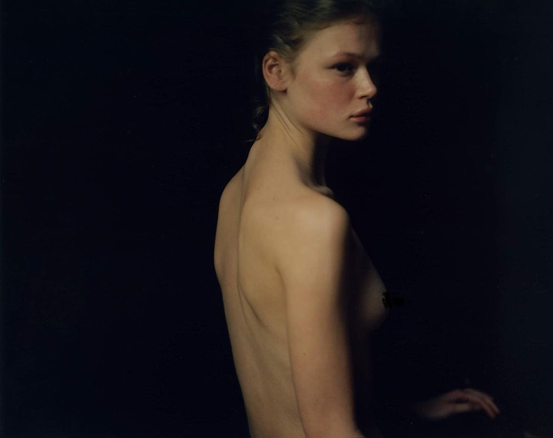 Striking studio portrait of a fair-skinned young woman with bare shoulders against a dark backdrop, illuminated by soft, directional light.