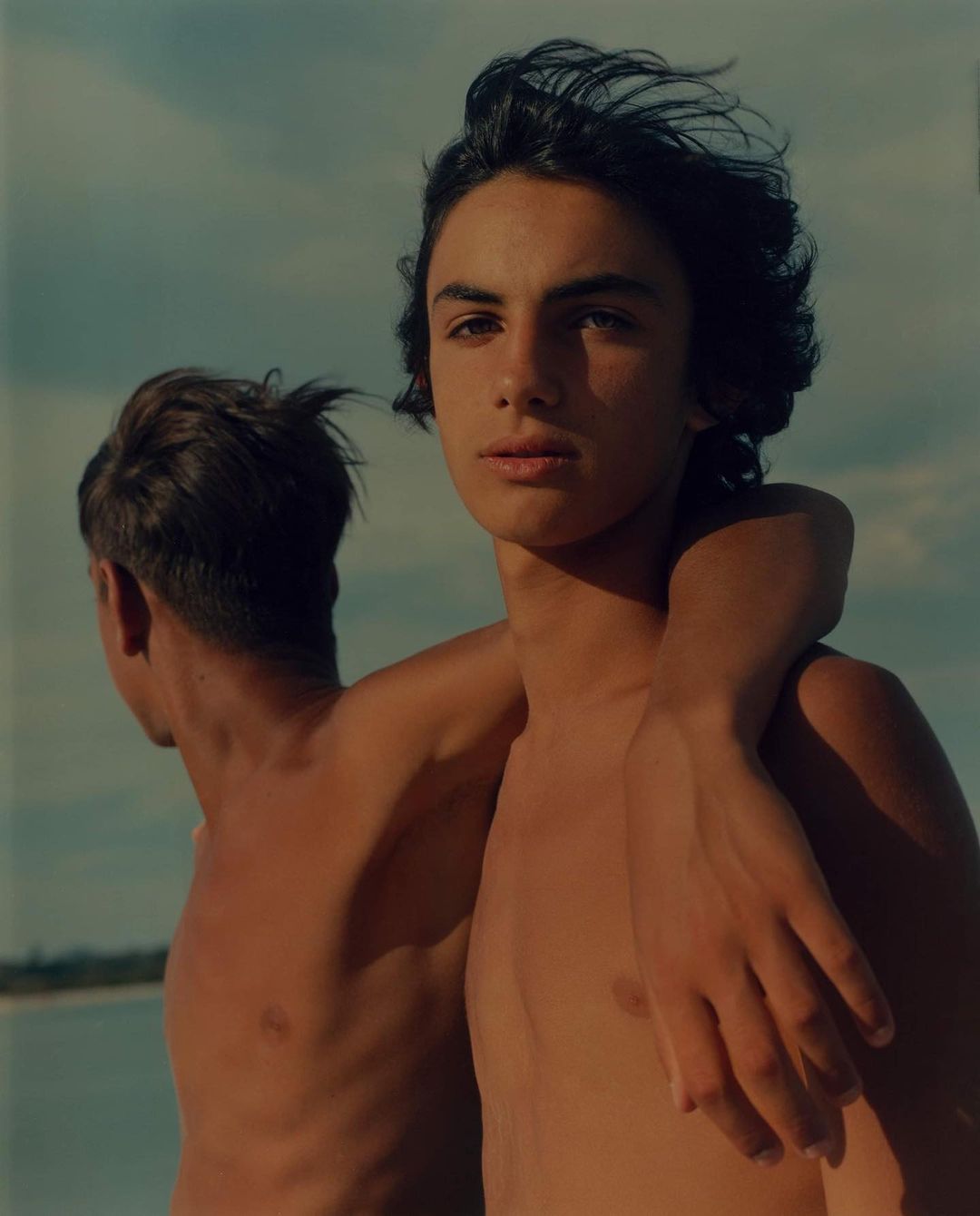 Two topless adult men stand close together outdoors against a pale sky. The primary subject faces forward with his arm around the other man.