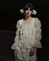 Model in white ruffled dress with sheer lace sleeves and matching hair bows against dark background.