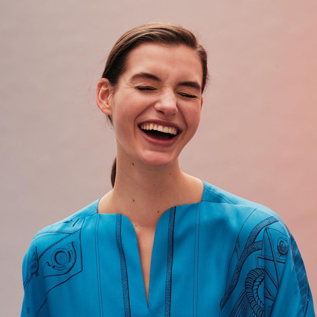 A happy, light-skinned adult woman smiles with teeth showing in a close up shot wearing a bright turquoise top with black details.