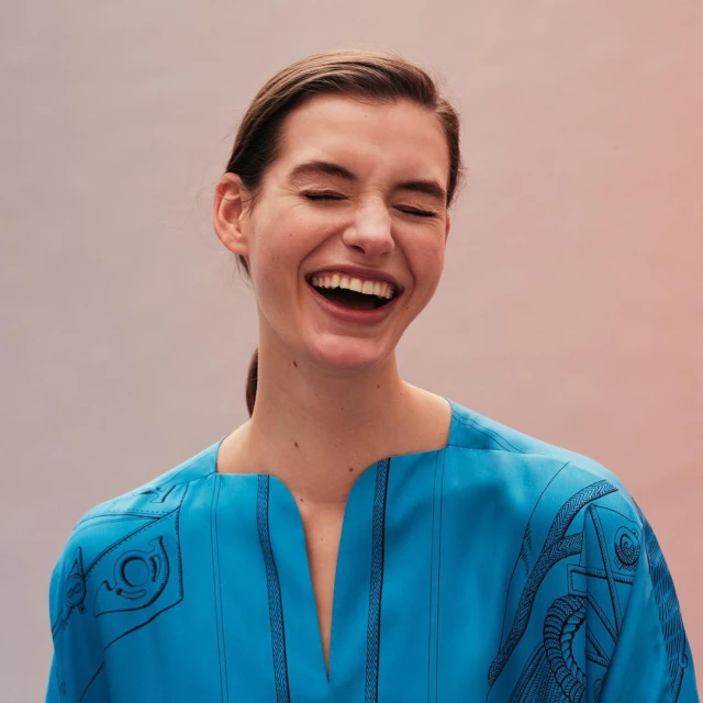 A happy, light-skinned adult woman smiles with teeth showing in a close up shot wearing a bright turquoise top with black details.