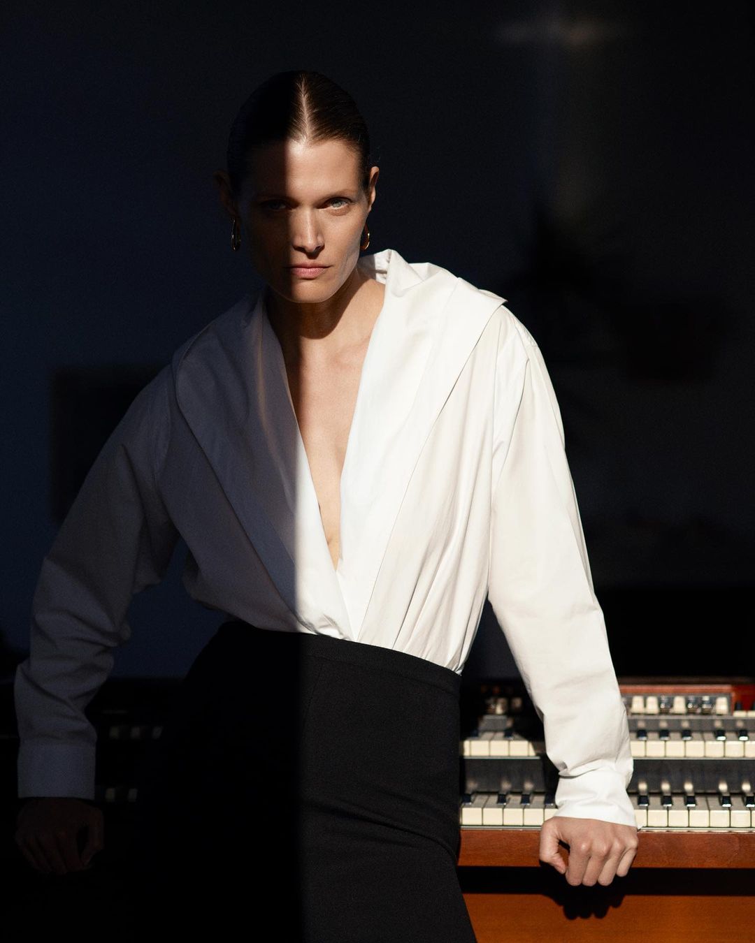 Indoor portrait of an adult woman in a white shirt and black skirt posing near a vintage organ with dramatic, contrasting lighting.