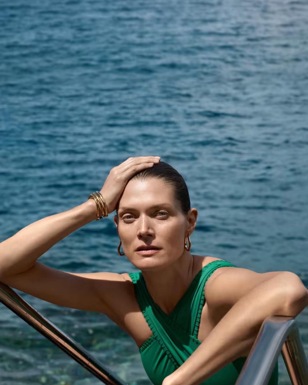 Woman in a green swimsuit poses by the ocean with her hand on her head, wearing gold jewelry and hoop earrings on a sunny day.